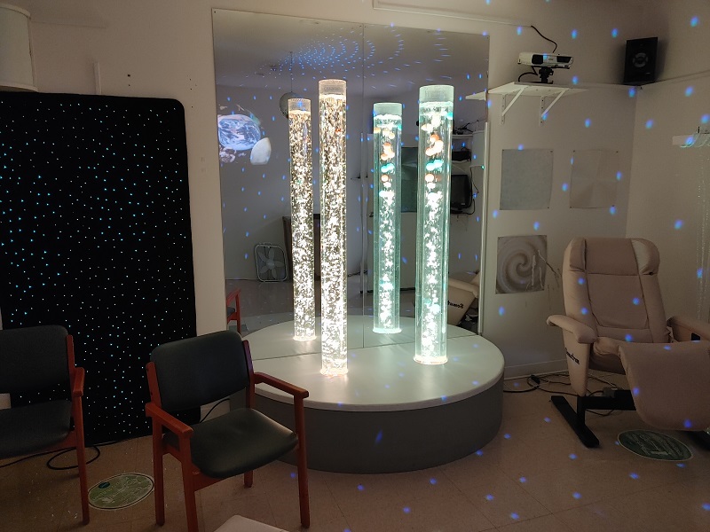 A "Snoezelen Room" with tall glittering tubes in front of a mirror. Blue dots shine on the walls from a projector. There are two empty chairs and a black wall panel covered in tiny dots of light. 
